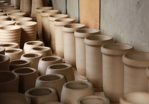 A row of unglazed, light beige ceramic planters with flared rims, arranged in rows on a floor inside a workshop or studio.