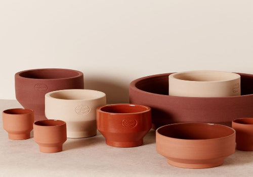 A set of terracotta and stoneware plant pots and cups in earthy terracotta, rust red, and cream beige colors, arranged on a neutral beige surface against a light beige background.